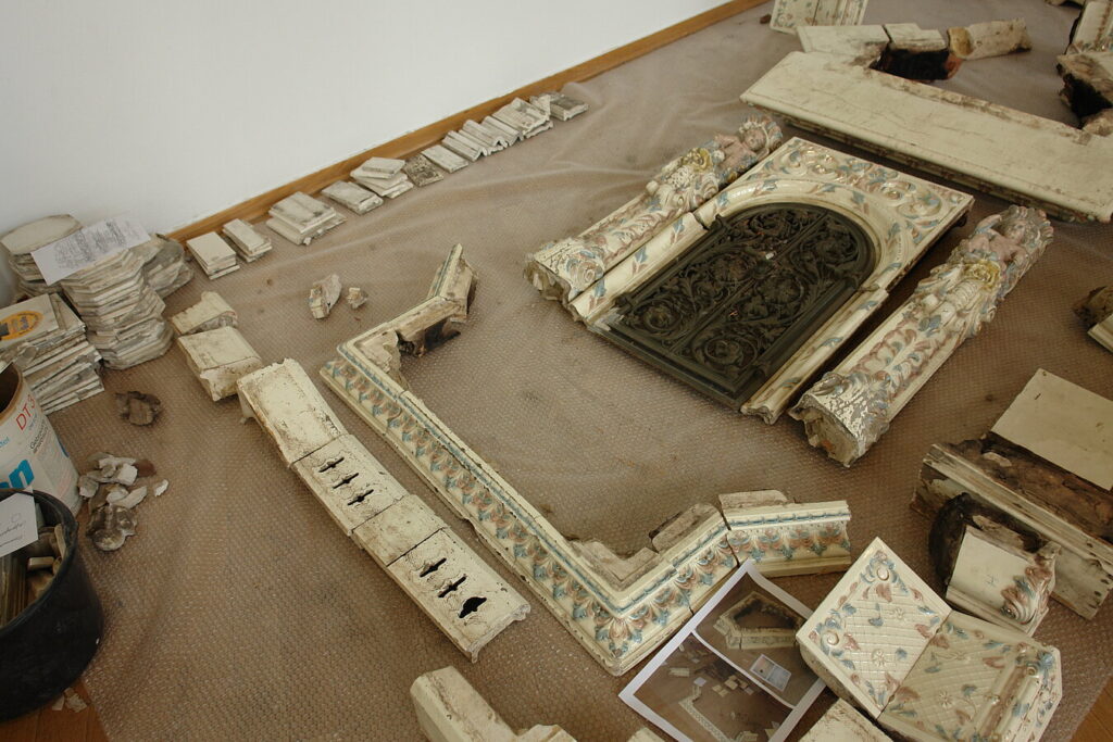 Zerlegte, verzierte Kaminteile aus Keramik liegen auf einem Boden, darunter Säulen und Rahmen mit floralem Muster, © Stadtmuseum Oldenburg