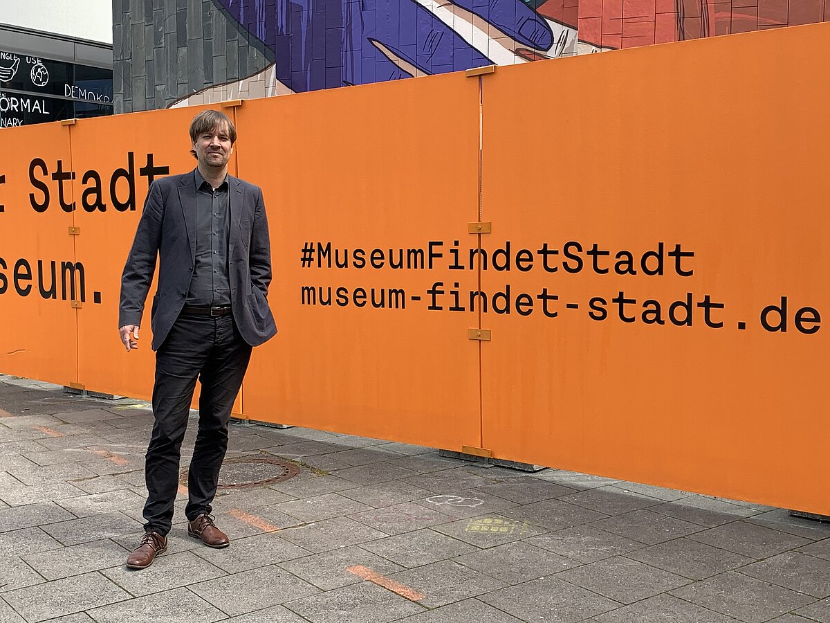 Pressebild Museum Findet Stadt 1 Foto Stadtmuseum Oldenburg Museumsleiter Dr. Steffen Wiegmann steht vor einer orangefarbenen Bauzaunwand mit dem Schriftzug '#MuseumFindetStadt museum-findet-stadt.de', © Stadtmuseum Oldenburg