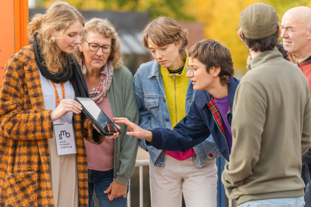 Eine Gruppe mit sechs Personen steht im Freien um ein Tablet herum und sieht sich darauf etwas an, © Studio 3x3