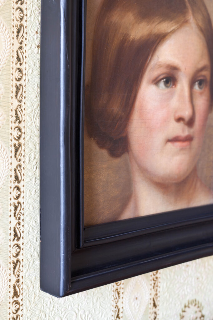 Gemälde aus dem Sammlungsbestand. Foto: Gerlinde Domininghaus Porträt einer Frau mit braunen Haaren, gerahmt in einem schwarzen Holzrahmen, an einer gemusterten Tapete hängend, © Gerlinde Domininghaus