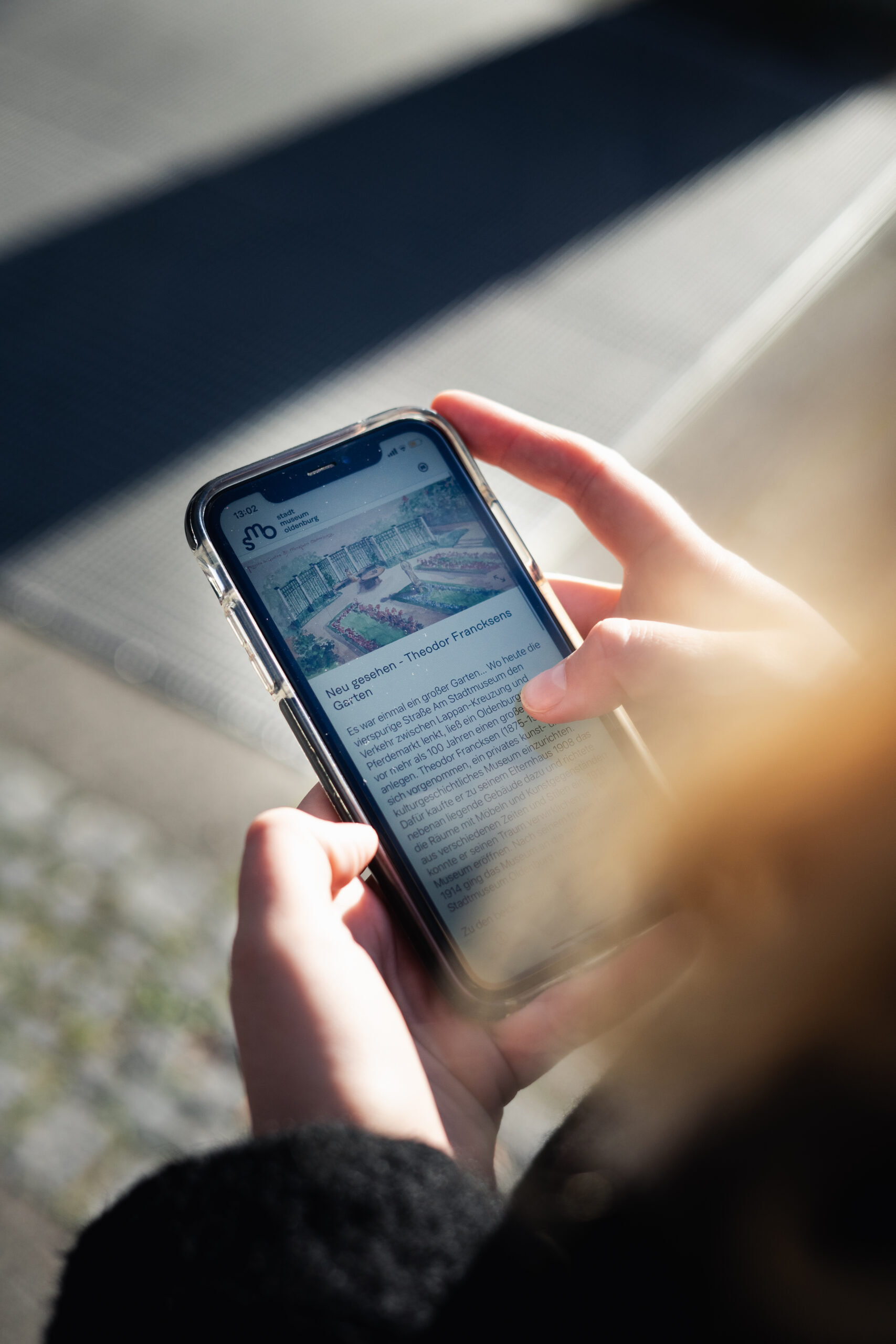 SMO Garten Neu Gesehen, © Stadtmuseum Oldenburg Smartphone wird gehalten und zeigt Einleitungstext über Theodor Francksens Garten ehemaligen am Stadtmuseum Oldenburg in der Garten-App, © Stadtmuseum Oldenburg