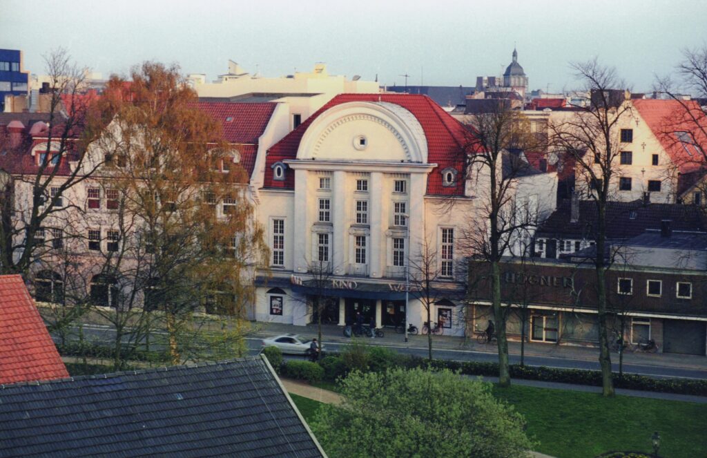 Abb. 8 BA 22466 Peter Kreier Wallkino Heiligengeiststr 2004 Historisches Kinogebäude mit rotem Mansarddach und weißer Fassade in einer Stadtumgebung, umgeben von Bäumen und weiteren Gebäuden