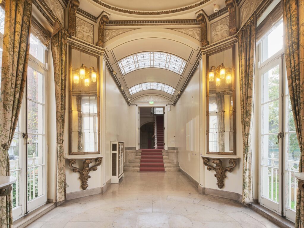 Heller, eleganter Flur in den historischen Villen mit großen Fenstern, zwei Wandspiegeln und einer Treppe mit rotem Teppich am Ende, © Caspar Sessler