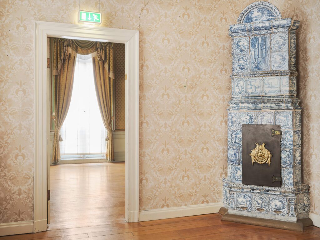 Heller Raum mit blau gekacheltem Kamin und Blick in den nächsten Raum, © Caspar Sessler