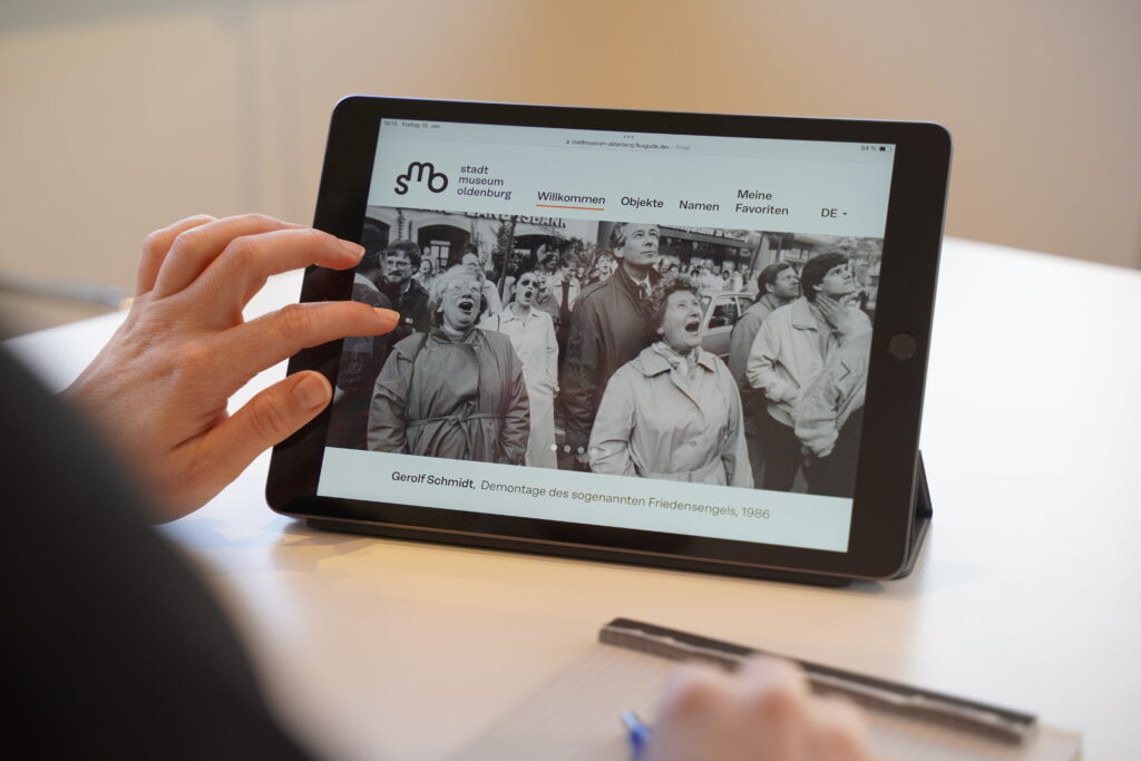 Die Online Sammlung des SMO auf dem Tablet, © Stadtmuseum Oldenburg Ein Tablet zeigt die Online-Sammlung des Stadtmuseums Oldenburg mit historischem Schwarz-Weiß-Foto von Demonstranten 1986, © Stadtmuseum Oldenburg