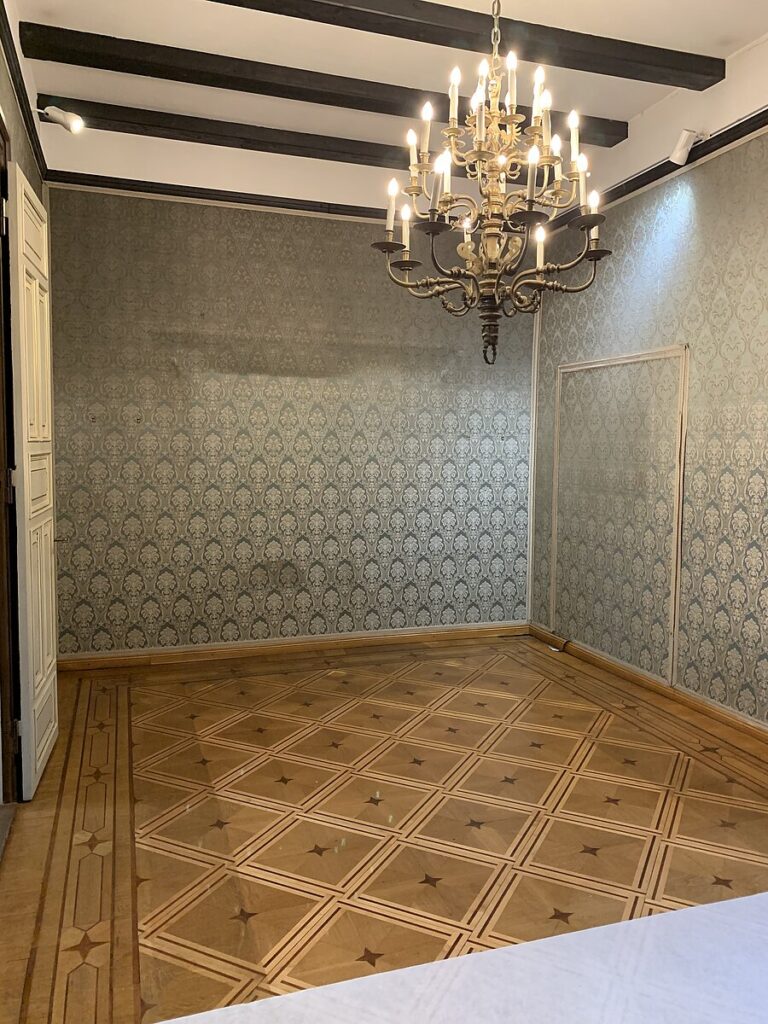 Leerer Raum der historischen Villen mit gemusterten Tapeten, Parkettboden mit Sternmotiven und einem Kronleuchter an der Decke, © Stadtmuseum Oldenburg