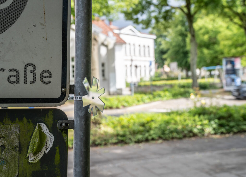 Kleiner sternchen-förmiger Spiegel an einem Straßenschild angebracht, im Hintergrund Bürgersteig mit Begrünung und Gebäude, © Stephan Walzl