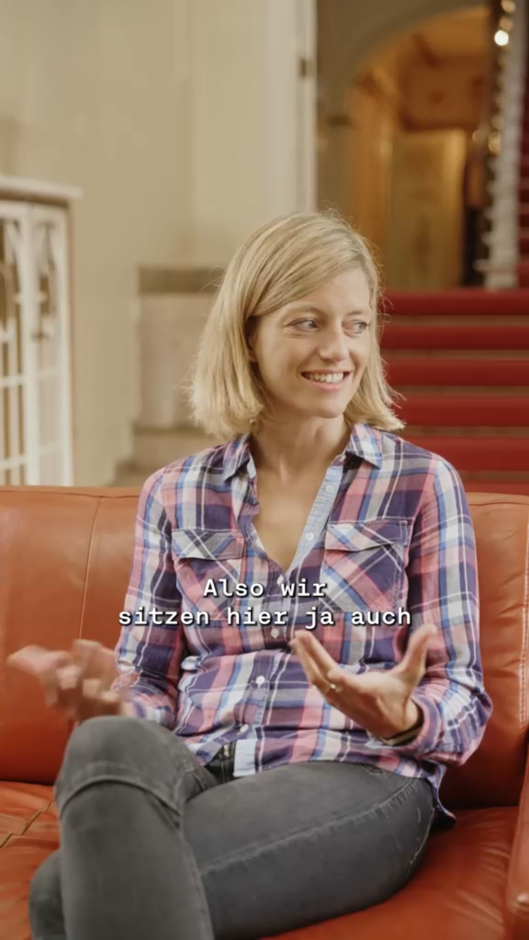 Jüngere Person sitzt auf rot-orangenem Sofa, im Hintergrund Flur der Francksenvilla mit rotem Teppich, © Stadtmuseum Oldenburg