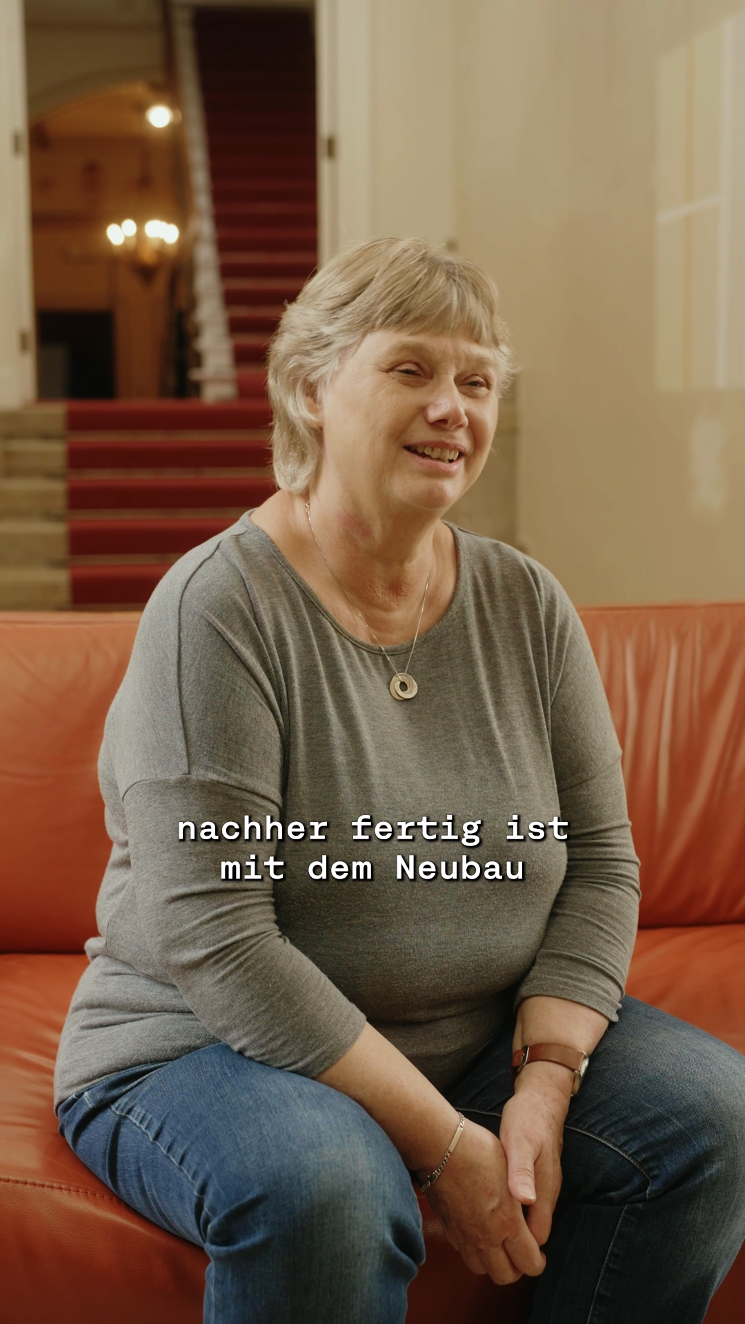 Ältere Person sitzt auf rot-orangenem Sofa, im Hintergrund Flur der Francksenvilla mit rotem Teppich, © Stadtmuseum Oldenburg