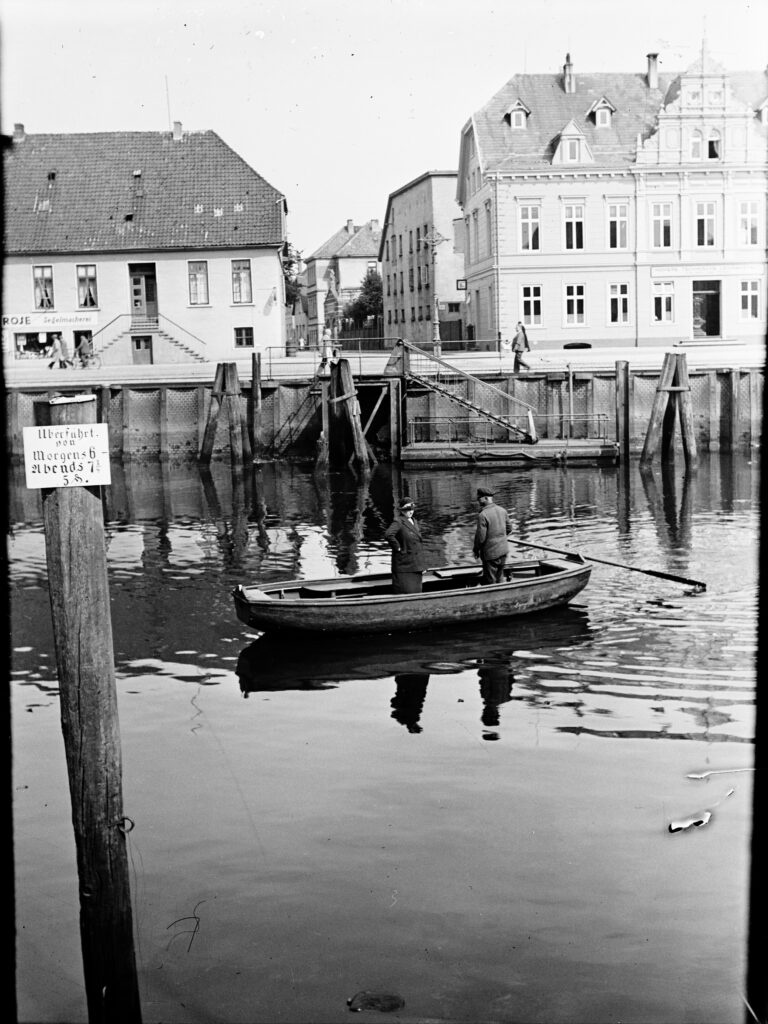 Ein kleines Boot auf dem Wasser, darauf zwei Personen, im Hintergrund eine Häuserzeile und die Befestigung des Hafens, © Stadtmuseum Oldenburg