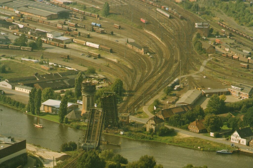 Vehrere Eisenbahnschienen laufen auf eine hochgezogene Bahnbrücke über einen Fluss zu, neben der Brücke ein Wasserturm, © Stadtmuseum Oldenburg