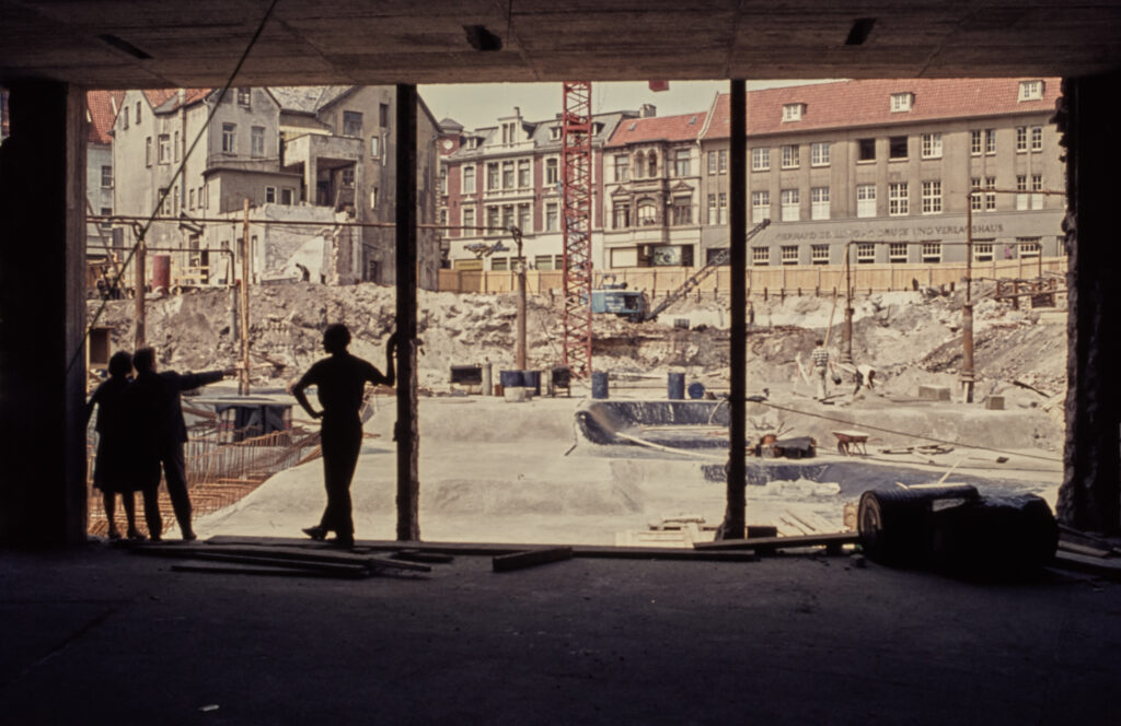 Personen stehen in einem abgebrochenen Gebäude und blicken auf eine Baustelle nach draußen, dort zu sehen Schutt und Werkzeuge, © Stadtmuseum Oldenburg/Sammlung Willy Schröder