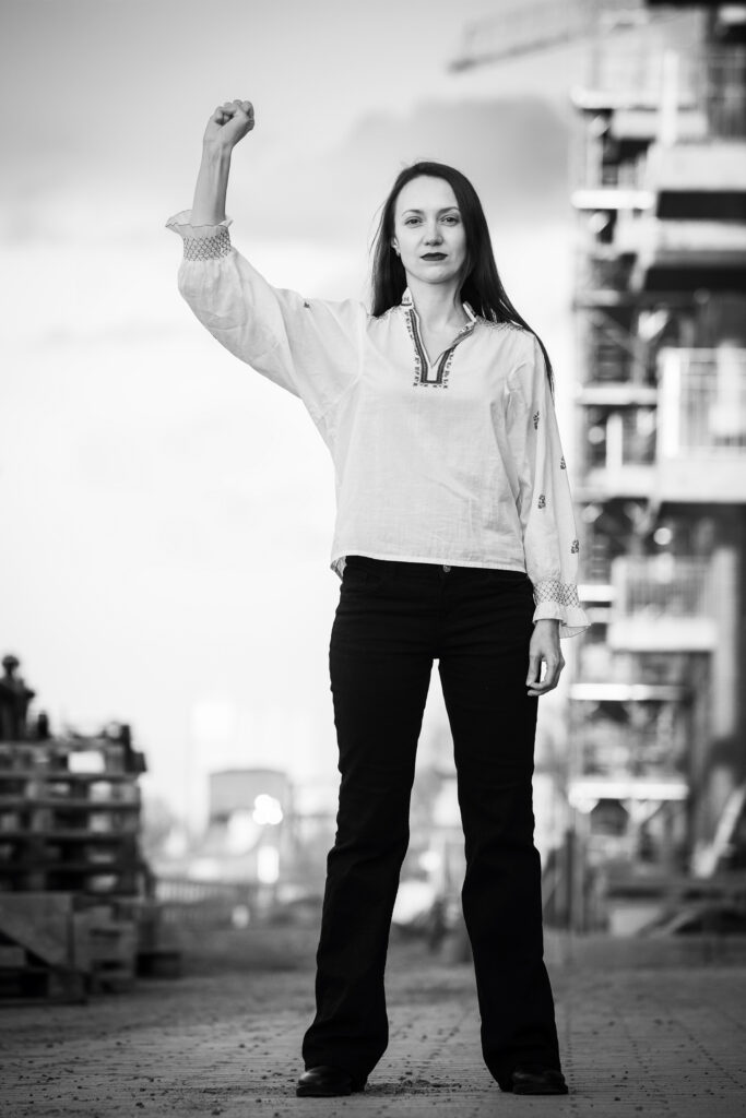 Antonina steht mit erhobener Faust in traditioneller weißer Bluse und schwarzer Hose vor einer Baustelle am Oldenburger Hafen, © Gerlinde Domininghaus