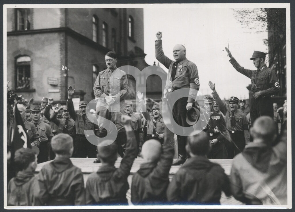 Abb 2 BA 10809 Hitler Am Pferdemarkt Wasserzeichen Eine große Gruppe Personen steht in Uniformen um einen Bühnenaufbau und zeigt den Hitlergruß, auf der Bühne weitere Personen, mit Armbinden, darunter auch Adolf Hitler, © Stadtmuseum Oldenburg/Heinz Besser
