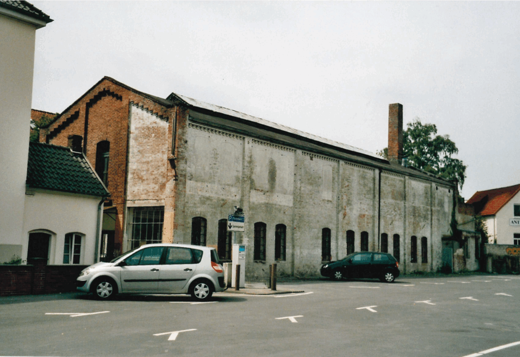 Seitenansicht der Halle vom Parkplatz, 03.06.2005, © Schlossmuseum Jever/Friedrich Precht Zwei Autos stehen auf einem Parkplatz vor einem länglichen Gebäude, mit rotem Backstein, eine Wand ist weiß gestrichen, © Schlossmuseum Jever/Friedrich Precht