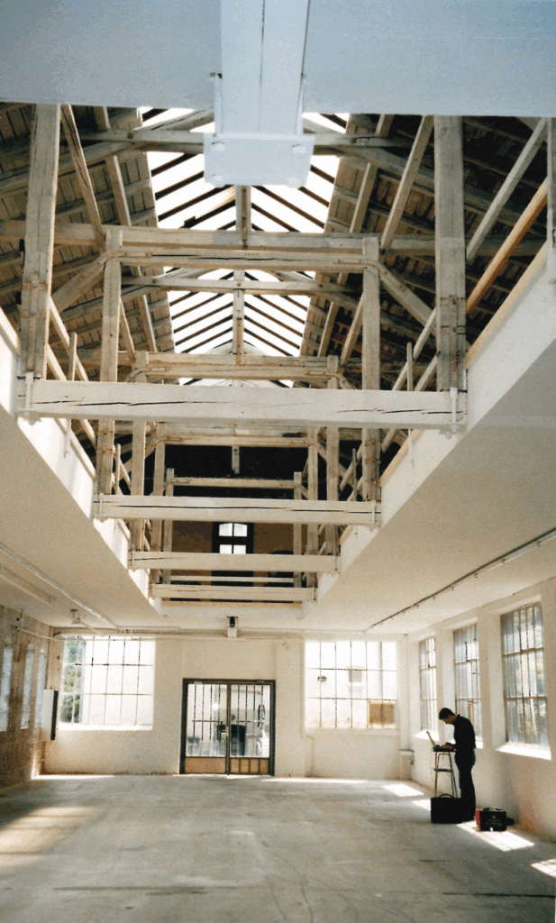 Innenansicht noch ohne Stahlkonstruktion, die 2012/2013 zur statischen Ertüchtigung der Halle eingebaut wurde, 03.06.2005, © Schlossmuseum Jever/Friedrich Precht Eine helle Halle mit Betonboden und vielen Fenstern, die Decke geöffnet zum oberen Stockwerk mit Holzbalken, ein Mann steht in der Halle an einem Laptop, © Schlossmuseum Jever/Friedrich Precht