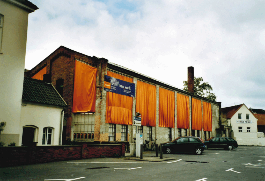 Stoffverhängung der Halle durch das Forum für Baukultur (bau_werk), um auf dessen Veranstaltungsprogramm und das Gebäude aufmerksam zu machen, 21.05.2006, © Schlossmuseum Jever/Friedrich Precht Orangene Tücher hängen über der Außenwand eines länglichen Gebäudes, davor ein Parkplatz, © Schlossmuseum Jever/Friedrich Precht