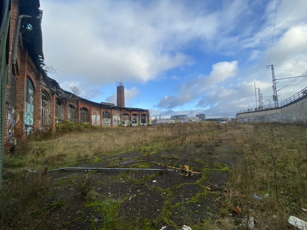 Abb. 10 Aktuell Verfällt Der Schuppen Zunehmend Und Die Fläche Davor Liegt Brach. 2022 Bild Privat Brachfläche mit Unkraut vor einer Backsteinhalle mit Toren, darauf Graffiti, © Privat