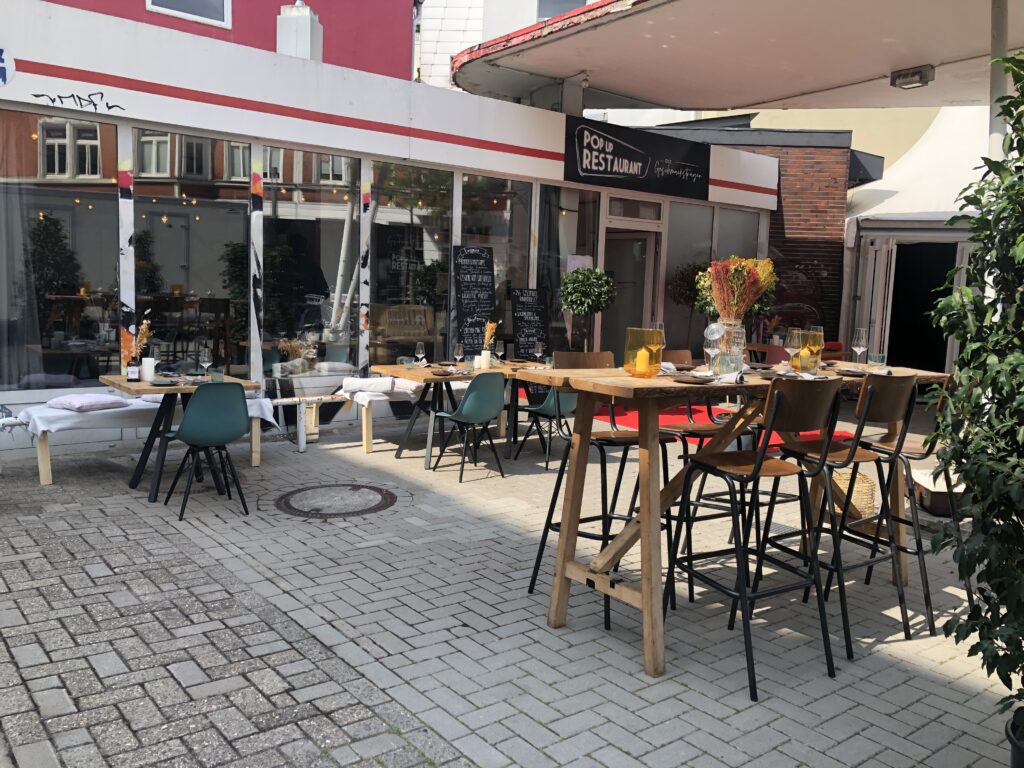 ehemaliges Tankstellengebäude mit großer Fensterfront, unter dem Vordach Tische und Stühle, © Kaiserküche