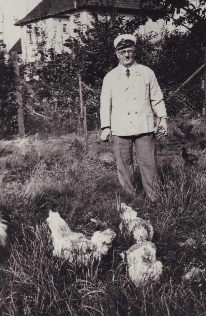 Mann mit weißer Jacke und Hut steht auf Rasen, vor ihm Hühner, hinter ihm ein Zaun, © Stadtmuseum Oldenburg/Slg. Friederichsen