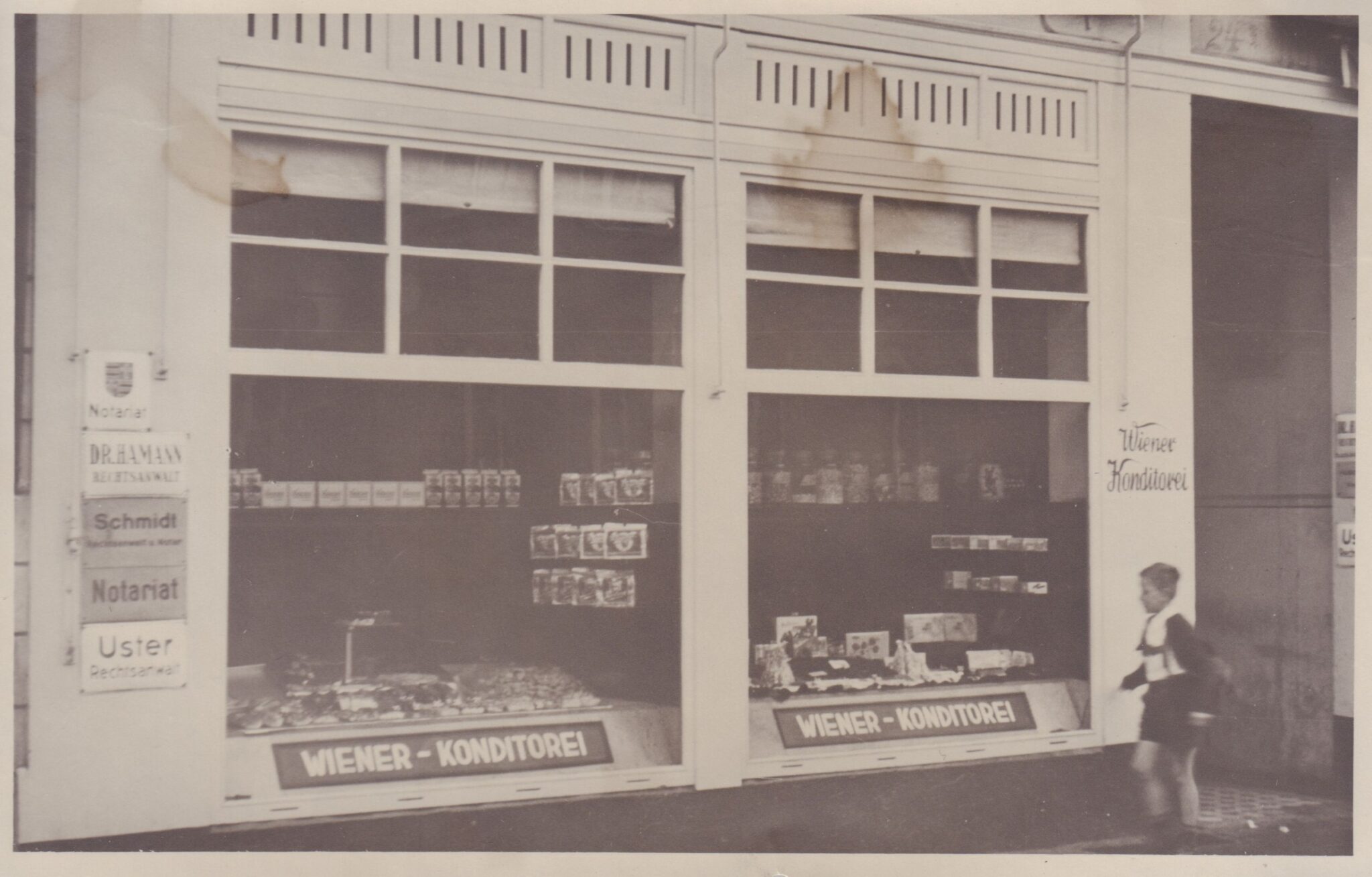 Abb. 2 Wiener Konditorei Um 1953 Schaufenster einer Konditorei in einem weißen Haus, davor rennt ein Kind, © Hans Janßen