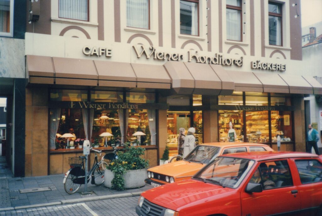 Abb. 3 Wiener Konditorei 1987 Ein rotes und ein orangenes Auto auf der Straße, dahinter ein Gebäude mit einer Konditorei und beleuchteten Schaufenstern, © Hans Janßen