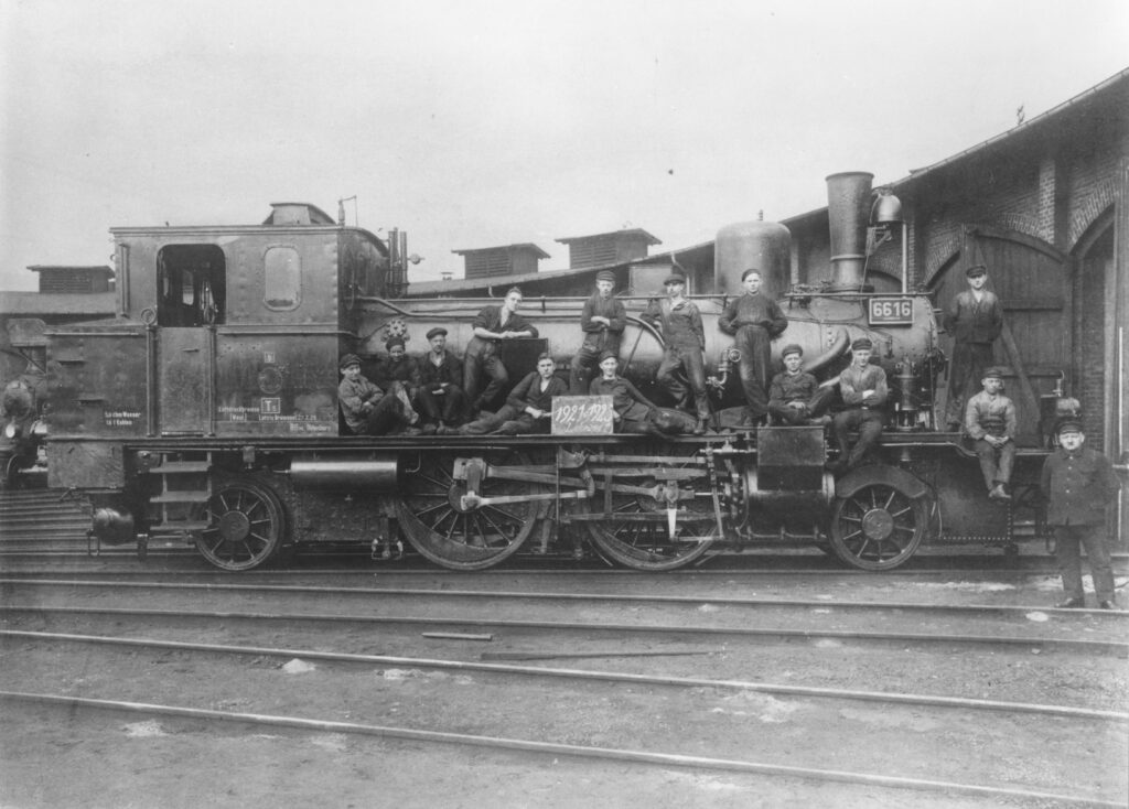 Abb. 4 Schlosserlehrlinge Vor Dem Ringlokschuppen1921 1925. Bild Stadtmuseum Oldenburg Eine Lokomotive auf Schienen, darauf sitzen und stehen verteilt Personen in schwarzer Arbeitskleidung, © Stadtmuseum Oldenburg