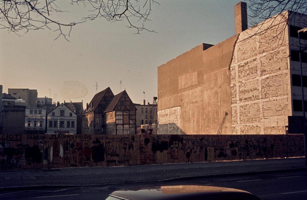 Abb. 5 Gebäude von der Sonne angeschienen, davor ein bemalter Bauzaun, im Hintergrund ein Fachwerkhaus, © Stadtmuseum Oldenburg, Slg. Willy Schröder