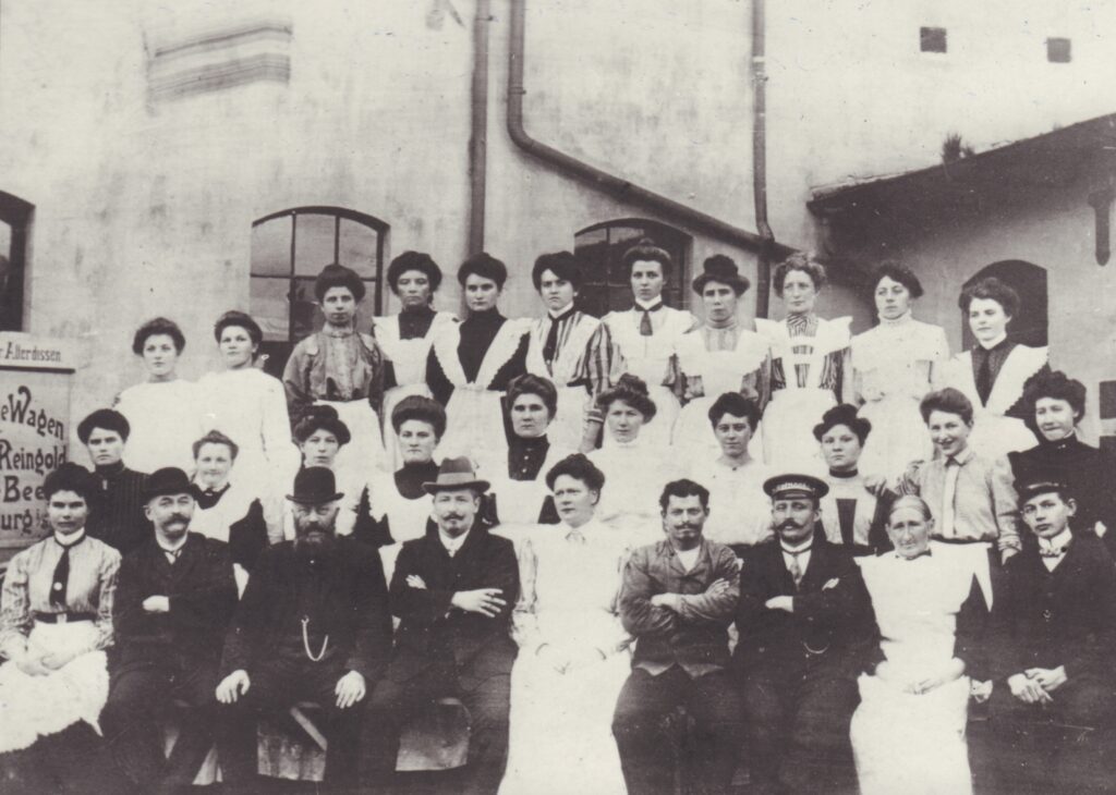 Mehrere Personen posieren sitzend und stehend für ein Foto, die Frauen mit weißen Schürzen, die Männer in dunklen Anzügen, vor einem weißen Haus, © Stadtmuseum Oldenburg/Slg. Friederichsen