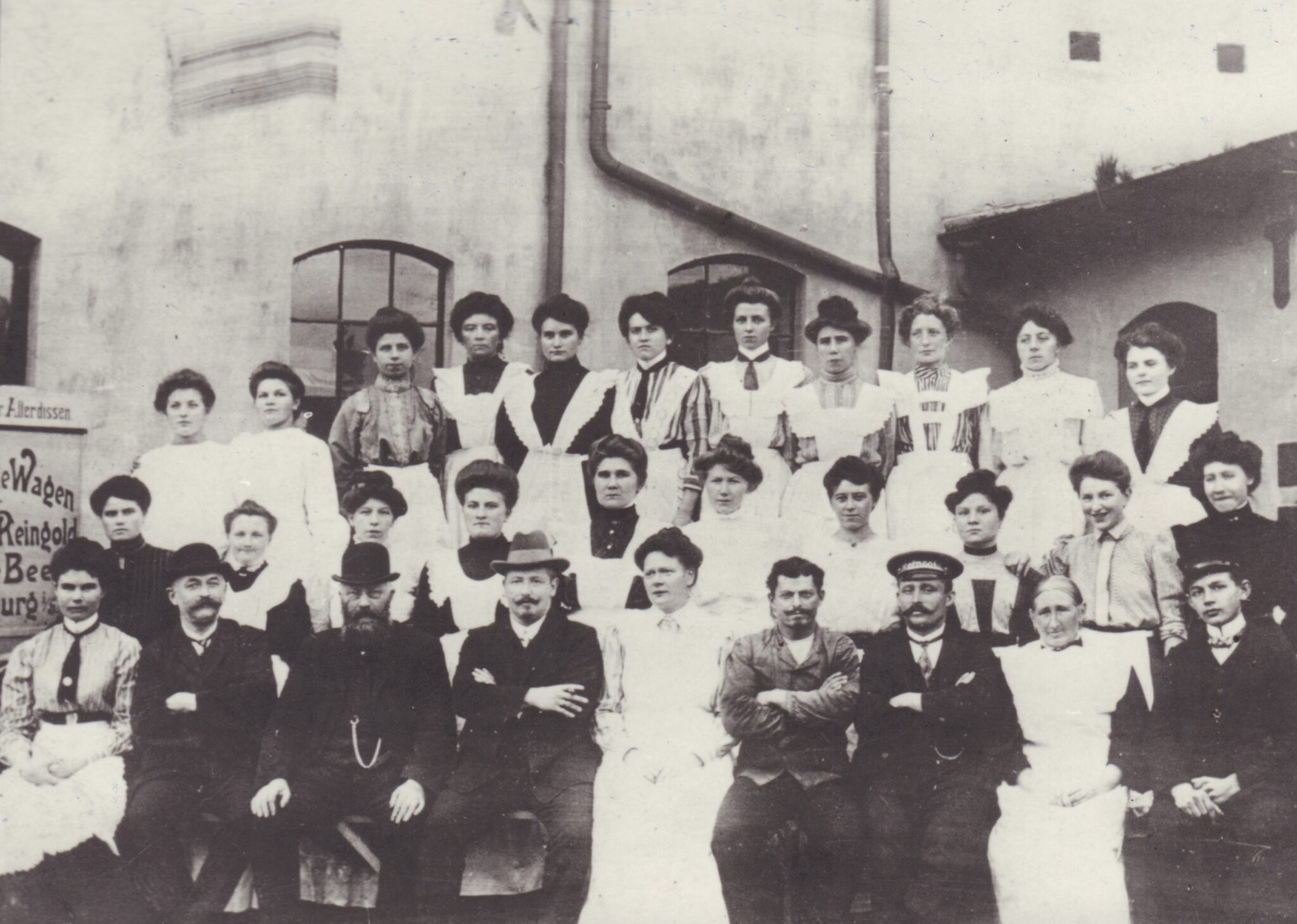 Ein Foto der bereits gewachsenen Belegschaft der Dampfwäscherei „Reingold“ im Jahre 1910. In der untersten Reihe sitzt Mathilde mittig im weißen Kleid, links daneben ist ihr Mann mit Hut und Schnäuzer, © Stadtmuseum Oldenburg/Slg. Friederichsen Mehrere Personen posieren sitzend und stehend für ein Foto, die Frauen mit weißen Schürzen, die Männer in dunklen Anzügen, vor einem weißen Haus, © Stadtmuseum Oldenburg/Slg. Friederichsen