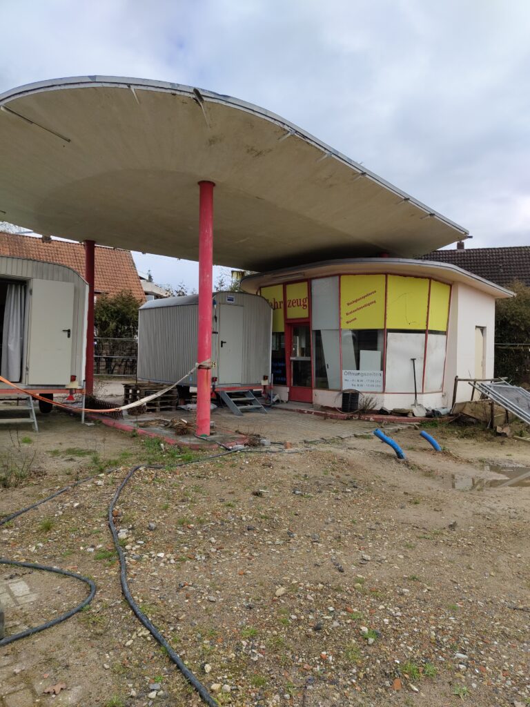 Stillgelegte kleine Tankstelle mit Vordach, blaue Rohre ragen aus der Erde