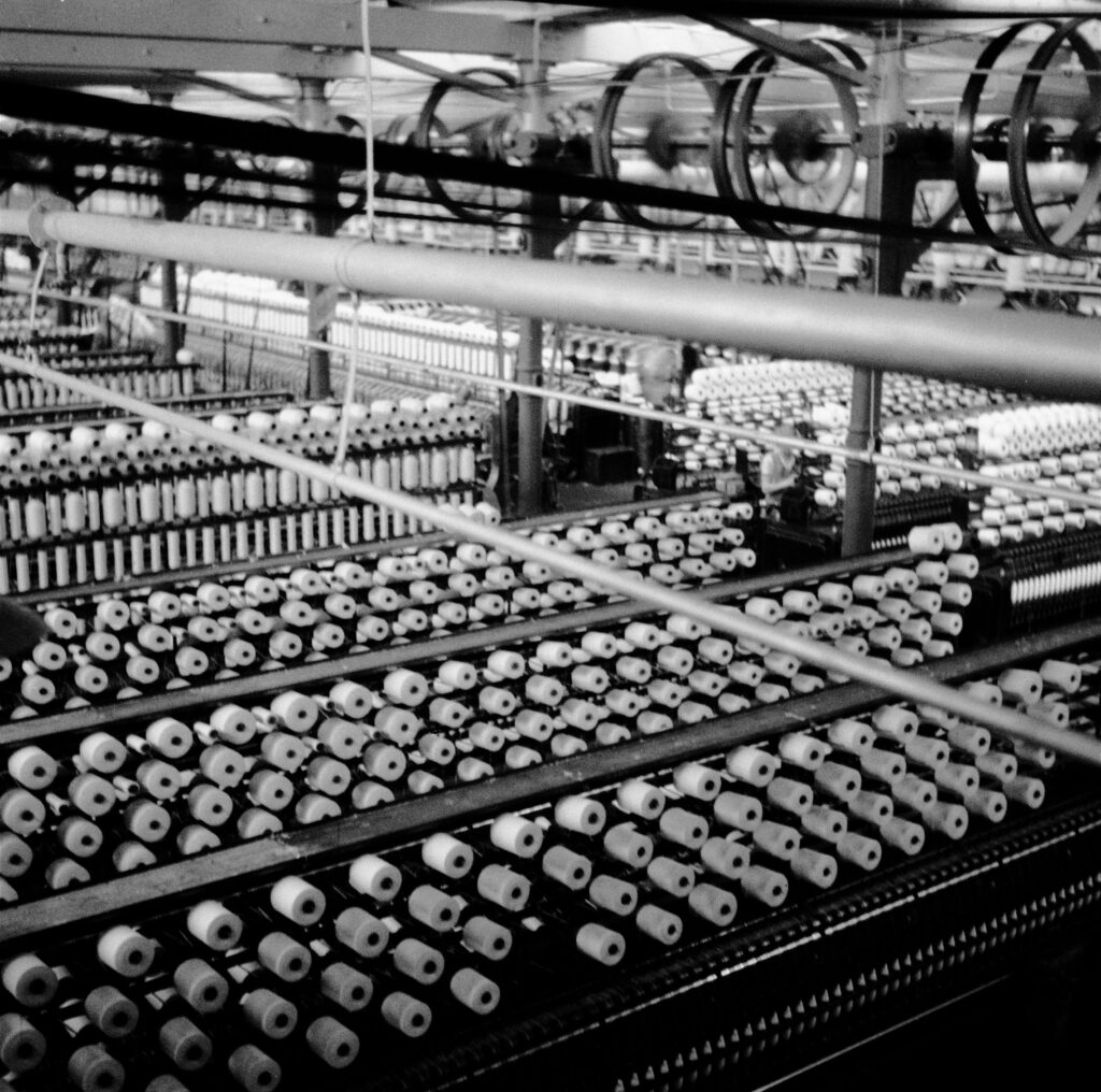 Gestelle gefüllt mit Fäden auf Spulen, in den Reihen arbeiten Personen, © Stadtmuseum Oldenburg, Kurt Wehlau.