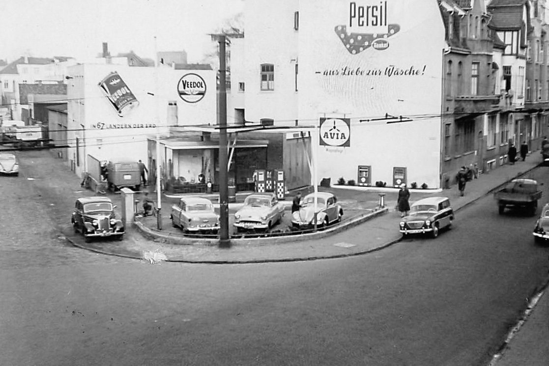 Abb. 6 Tankstelle Kaiserstraße 1950er Jahre © Firma Pickel Blick auf eine Tankstelle mit alten Autos, an den Gebäuden darum Produktwerbungen, © Firma Pickel