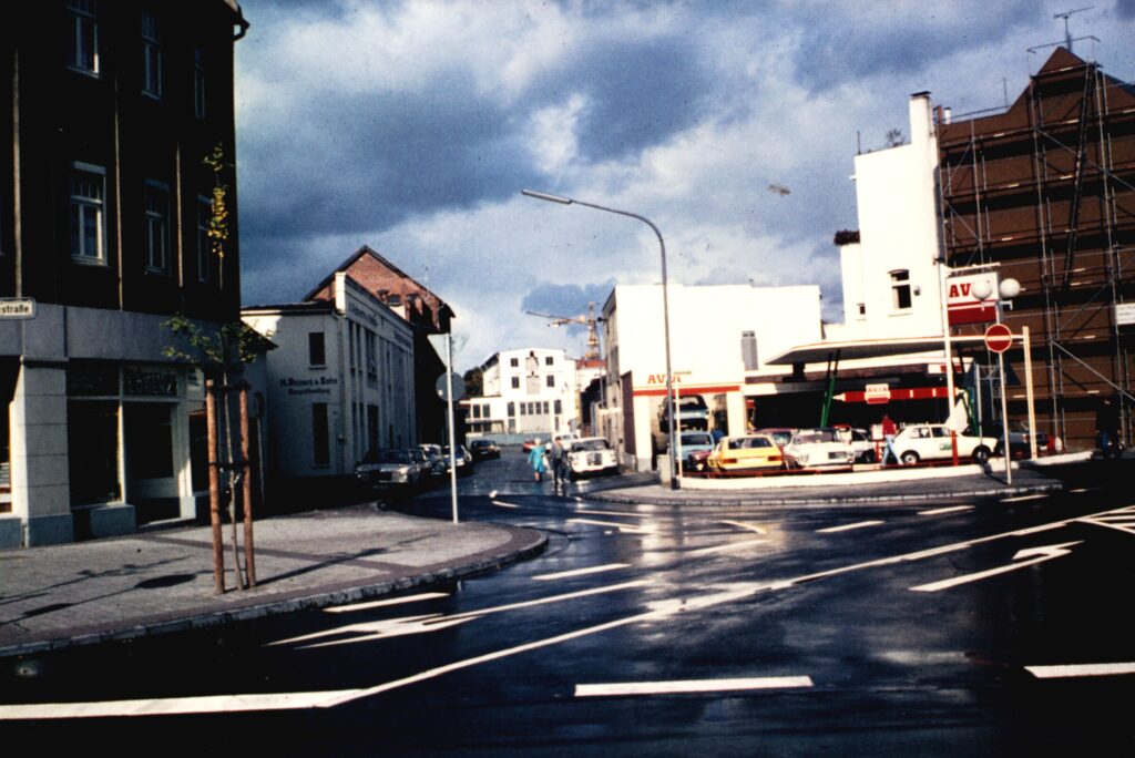 Straßenkreuzung mit hohen Gebäuden, hinten rechts eine kleine Tankstelle mit vielen Fahrzeugen