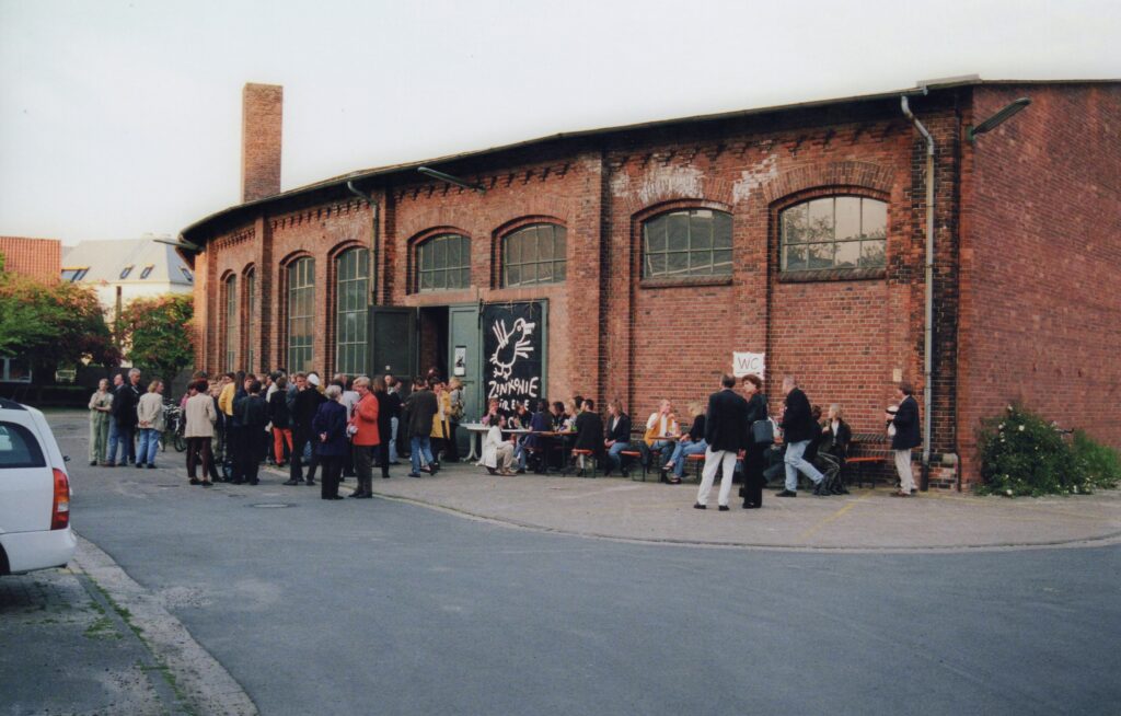 Abb. 7 Theateraufführung Im Leerstehenden Ringlokschuppen.1999. Bild Stadtmuseum Peter Kreier Personen versammelt vor einer Backsteinhalle, einige sitzen, unterhalten sich, © Stadtmuseum Oldenburg / Peter Kreier