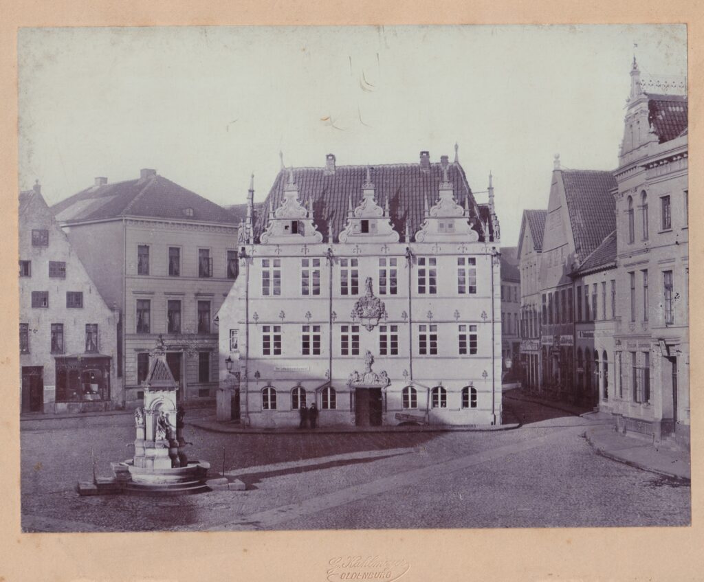 Abb.1 BA 1932 Marktplatz mit verziertem Brunnen in der Mitte dahinter ein reich verziertes Mehrstöckiges Gebäude, das Rathaus, © Stadtmuseum Oldenburg/Kahlmeyer