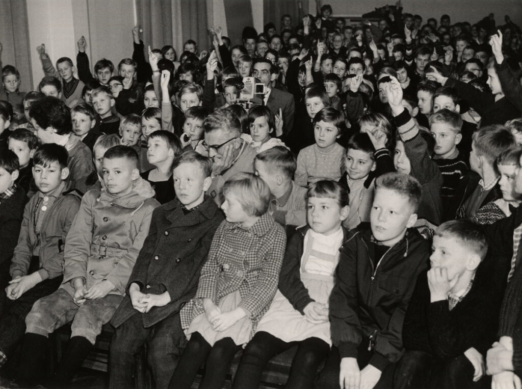 Viele Kinder sitzen in Reihen in einem Raum für einen Lichtbildvortrag, einige melden sich, © Stadtmuseum Oldenburg
