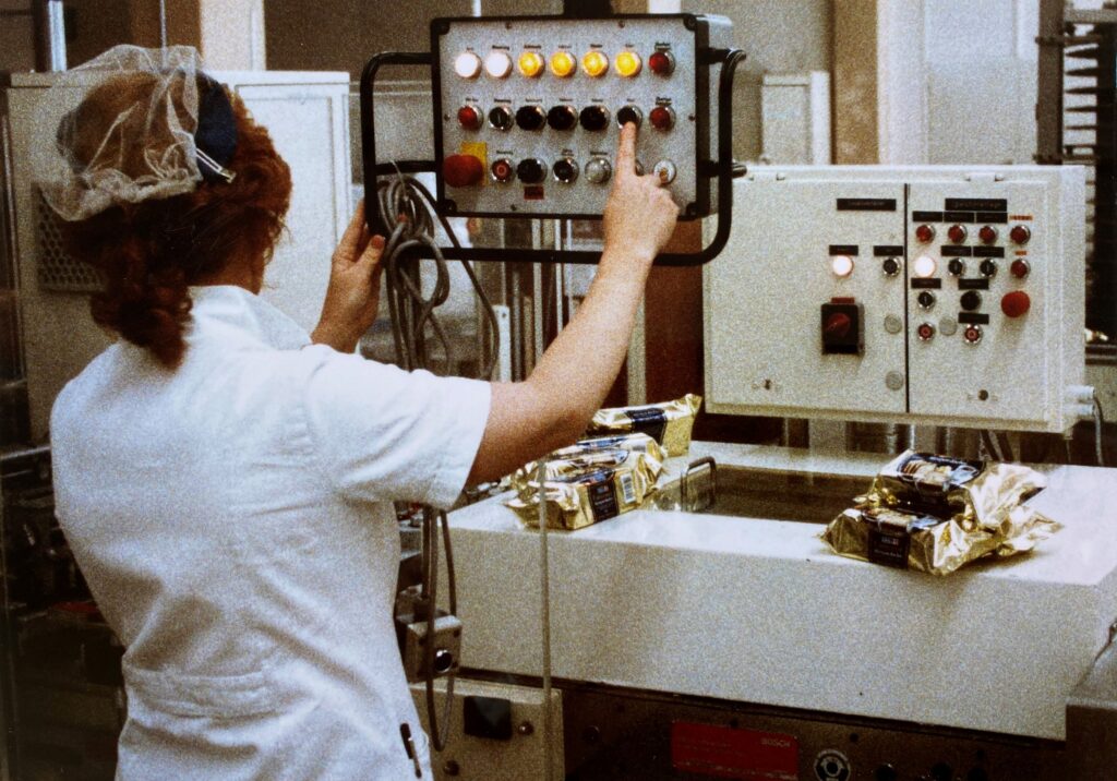 Eine Person in weißem Kittel mit Haarnetz bedient eine Maschine mit farbigen Knöpfen, darauf liegt abgepacktes Gebäck, © Bahlsen Gmbh & Co. KG