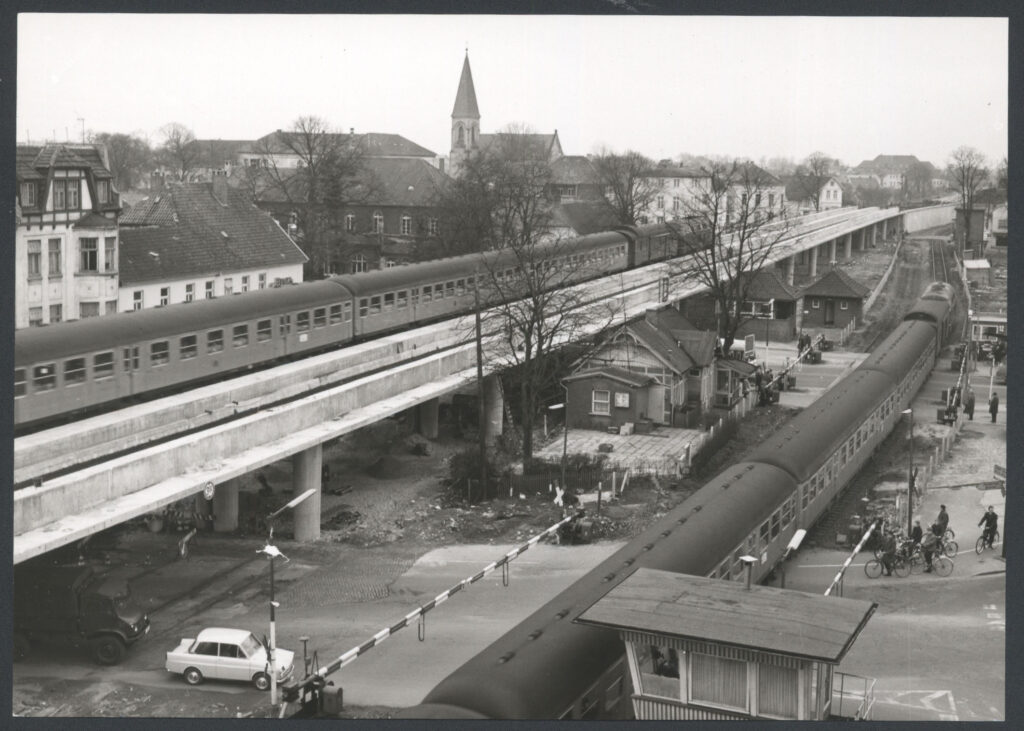 Abb.5 Ba 10711 Bahnhochlegung Am Pferdemarkt 1962 Blick von oben auf eine Bahnstrecke, auf der ein Zug fährt, Personen auf Fahrrädern und in Autos warten an der Schranke, am linken Rand eine Brücke auf der ein Zug fährt, © Stadtmuseum Oldenburg