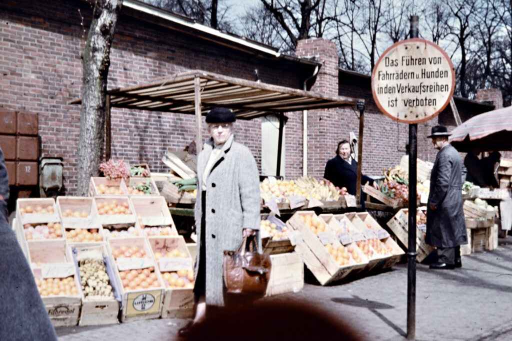 Abb.6 BA 27919 Pferdemarkt Ladenzeile Marktstand Um 1958 Käthe Nebel Gemüsekisten unter einem kleinen Unterstand, davor Personen beim Einkauf, © Stadtmuseum Oldenburg/Käthe Nebel