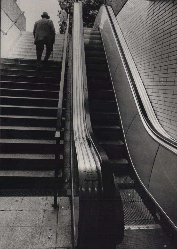 Aufgang einer Treppe, daneben eine Rolltreppe, eine Person mit Hut und Stock geht hinauf, © Schlossmuseum Jever/Jeddeloh