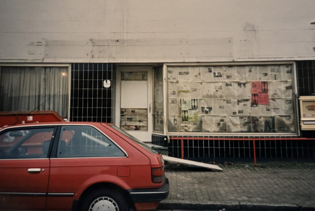 Rotes Auto vor einem Gebäude, dessen Schaufenster mit Zeitungen beklebt ist, © Mit freundlicher Genehmigung des „Na Und e. V.“.