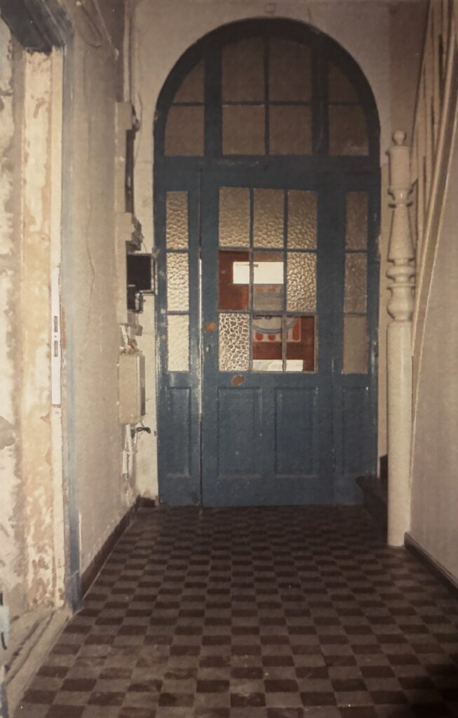 Blaue hohe Tür mit Glasfenstern und Bogen, davor gekachelter Boden in einem weißen Flur, © Mit freundlicher Genehmigung des „Na Und e. V.“.