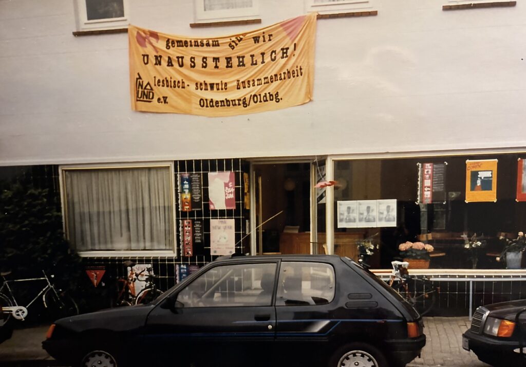 Schwarzes Auto vor einem weißen Haus mit schwarzen Kacheln am EIngang und einem Schaufenster, über dem Eingang gelbes Banner mit Aufschrift, © Mit freundlicher Genehmigung des „Na Und e. V.“.