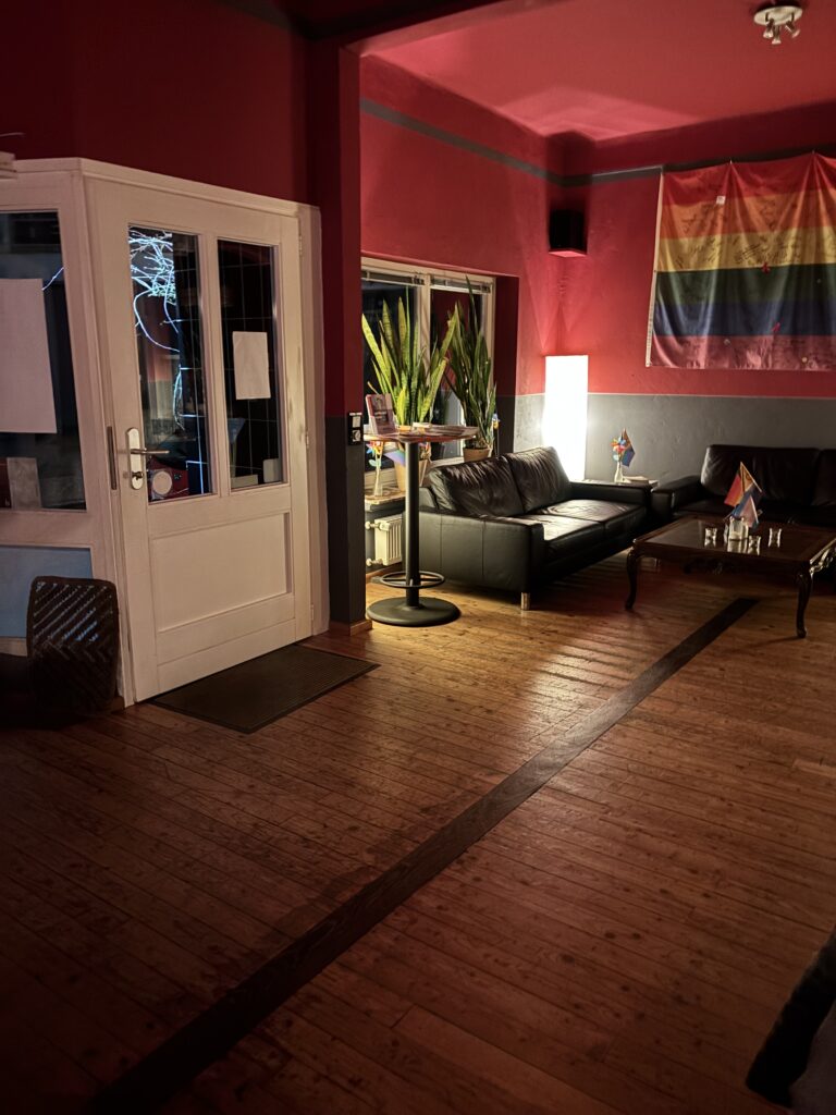 Schwach beleuchteter Raum mit Roter Wand und Sitzecke, an der Wand eine Regenbogenflagge, © Mit freundlicher Genehmigung des „Na Und e. V.“.