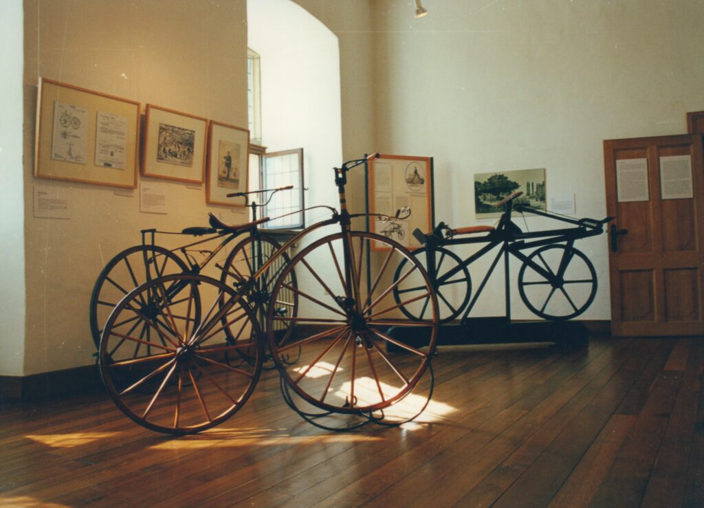 Ein Blick in die Dauerausstellung des Oldenburger Fahrradmuseums, © Stadtmuseum Oldenburg/Prov. Kalle Kalkhoff Historische Fahrräder stehen in einem Raum mit Parkettboden, an den Wänden Rahmen mit Bildern zu Fahrrädern, © Stadtmuseum Oldenburg/Prov. Kalle Kalkhoff