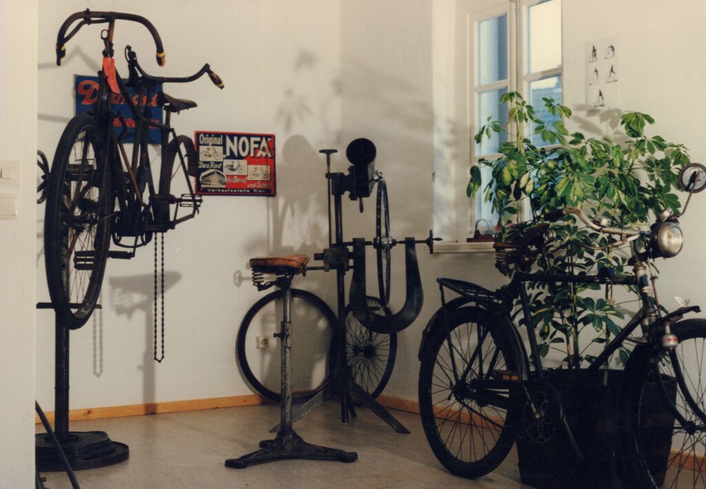 Ein Blick in die Dauerausstellung des Oldenburger Fahrradmuseums, © Stadtmuseum Oldenburg/Prov. Kalle Kalkhoff An Gestelle montierte Fahrräder in einer Ecke eines Raums, im Vordergrund eine Grünpflanze, © Stadtmuseum Oldenburg/Prov. Kalle Kalkhoff