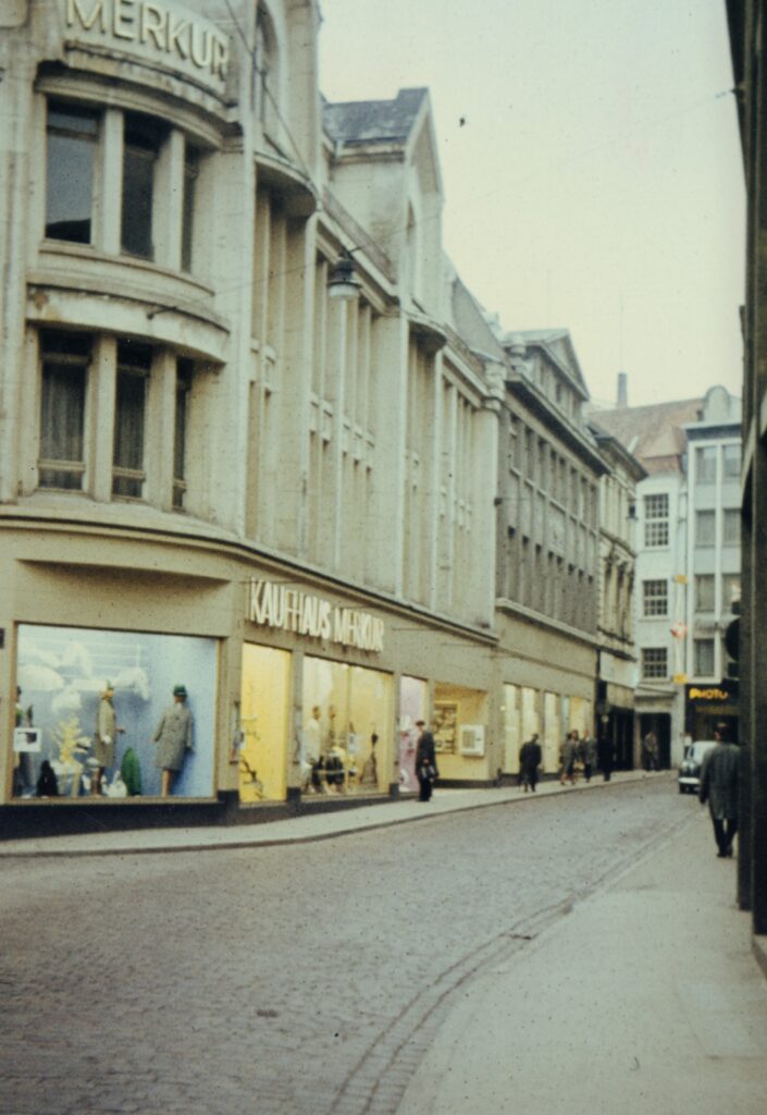 Häuserzeile an einer Straße mit Backsteinpflaster, mit großen Schaufenstern, am vorderen Gebäude die Aufschrift "Kaufhaus Merkur", © Stadtmuseum Oldenburg/Sammlung Schäfer