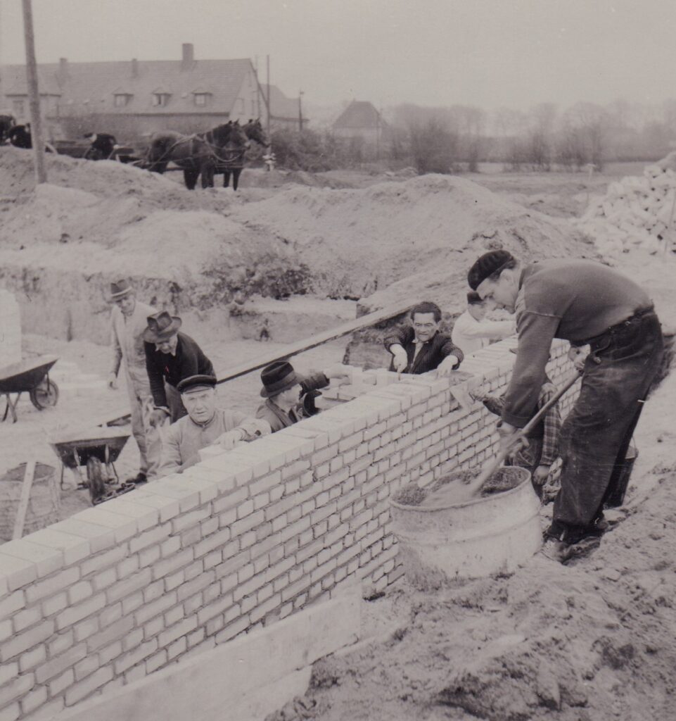 Männer arbeiten an einer Backsteinmauer, drum herum Sandberge, im Hintergrund eine Pferdekutsche, © Günter Nordhausen.