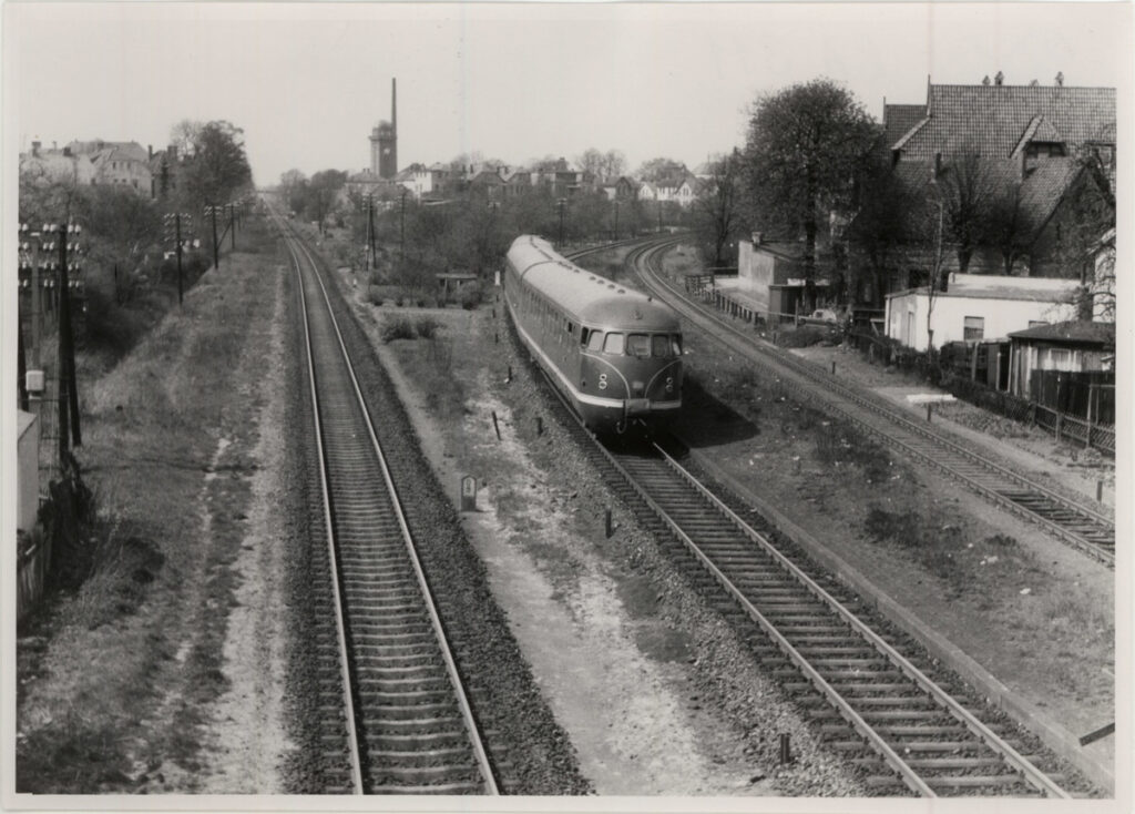 Die Bahnstrecken nach Leer und Wilhelmshaven fotografiert von der ehemaligen Fußgängerbrücke Ziegelhofstraße um 1963, © Stadtmuseum Oldenburg Bahnstrecke mit Gleisen, auf der ein alter Zug entlang fährt, © Stadtmuseum Oldenburg